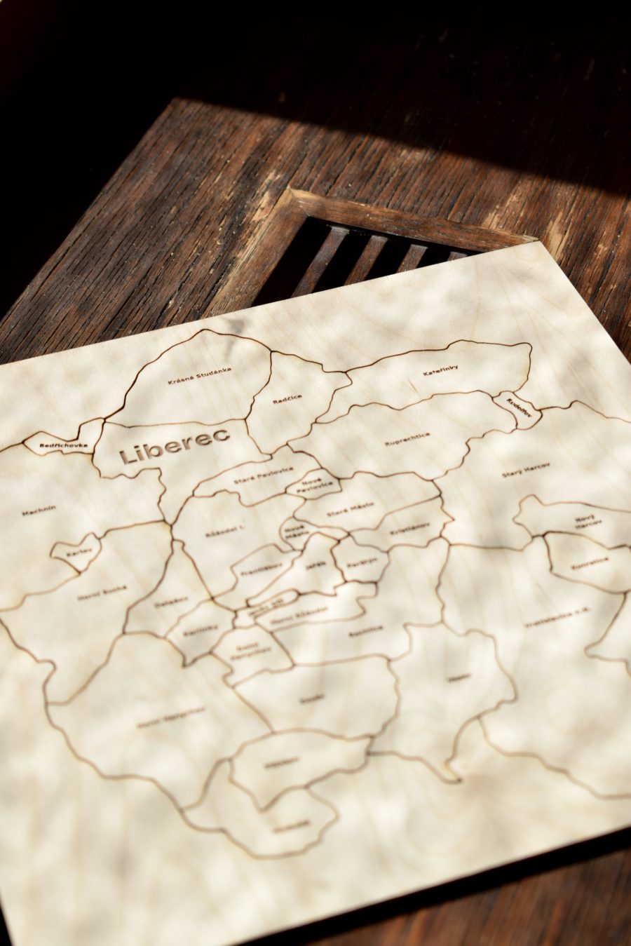 Dřevěné puzzle Liberce ležící na tmavé dřevěné lavici, v překližce jsou vypálené hranice a názvy všech 33 čtvrtí.